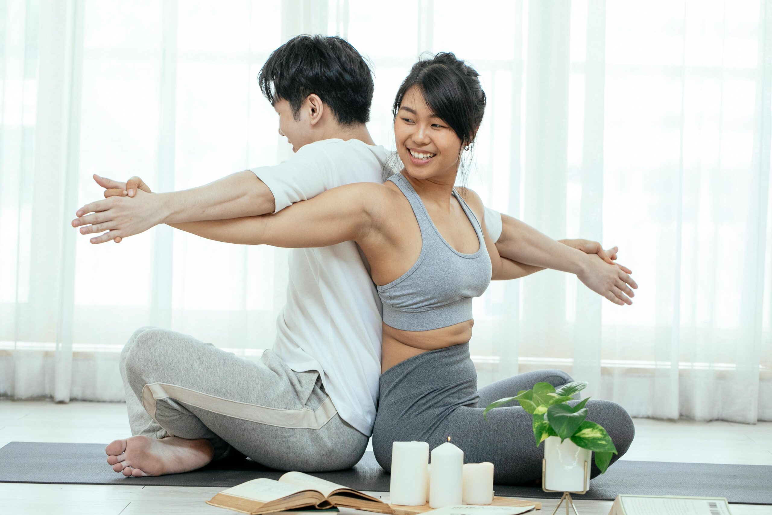 Smiling young Asian couple doing acro yoga while sitting back to back with crossed arms on mat near window