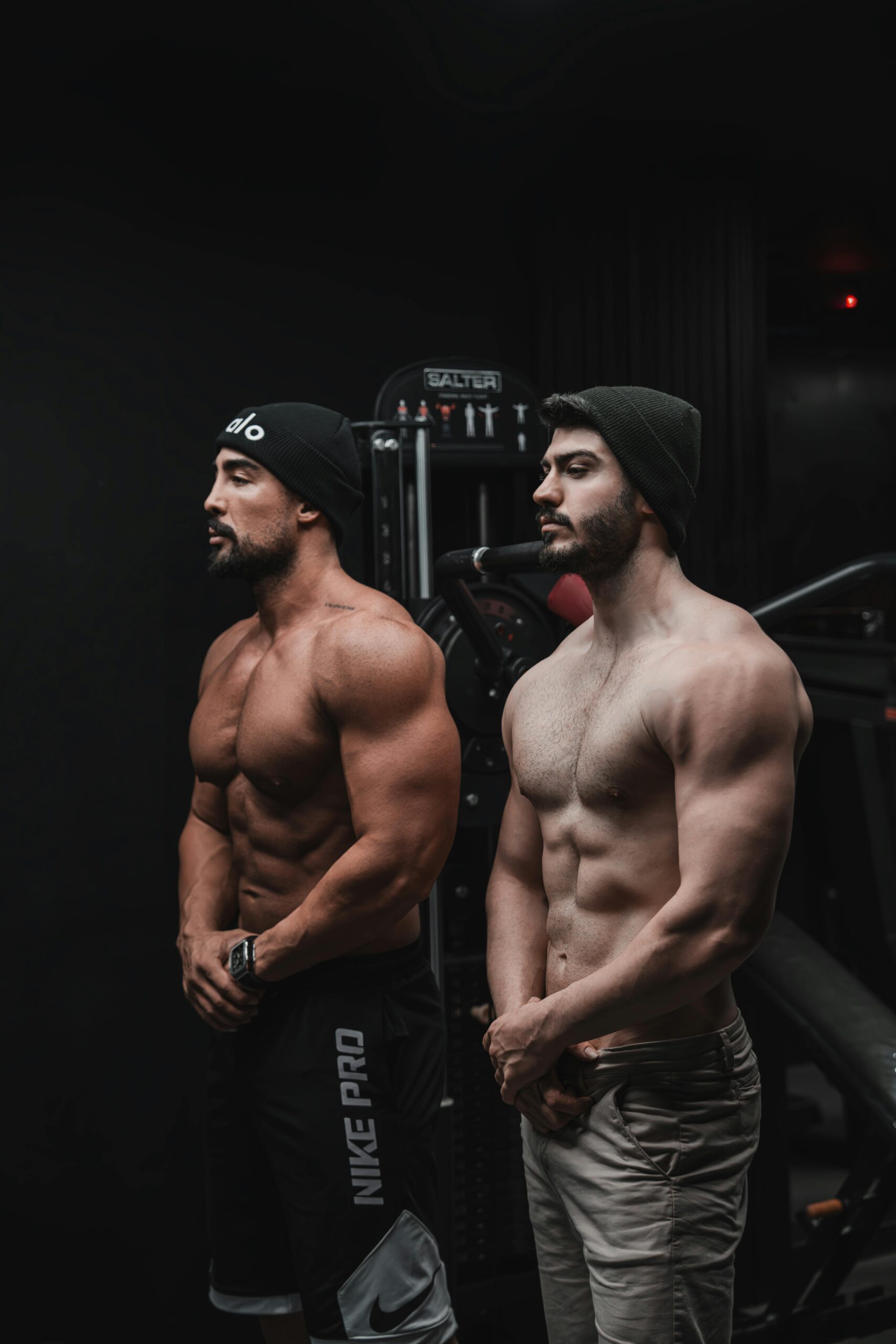 Two muscular men posing in a dimly lit gym, showcasing fitness and strength.