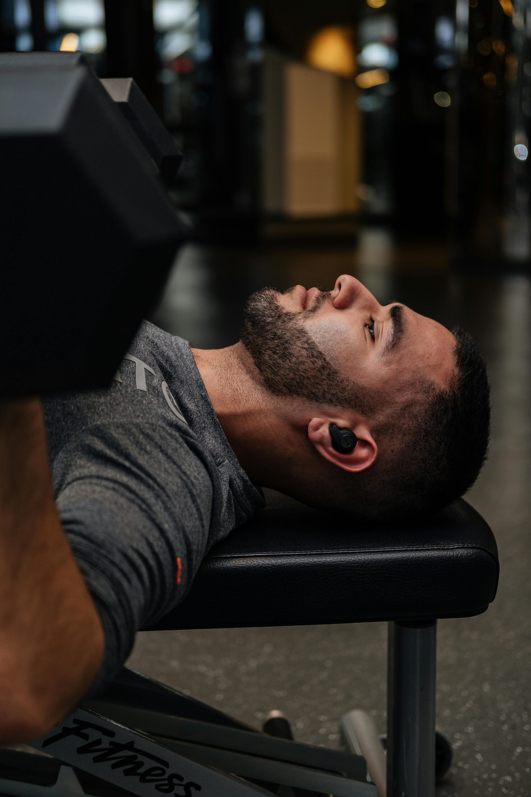 A focused man performing a bench press with dumbbells in a gym setting, wearing earbuds.