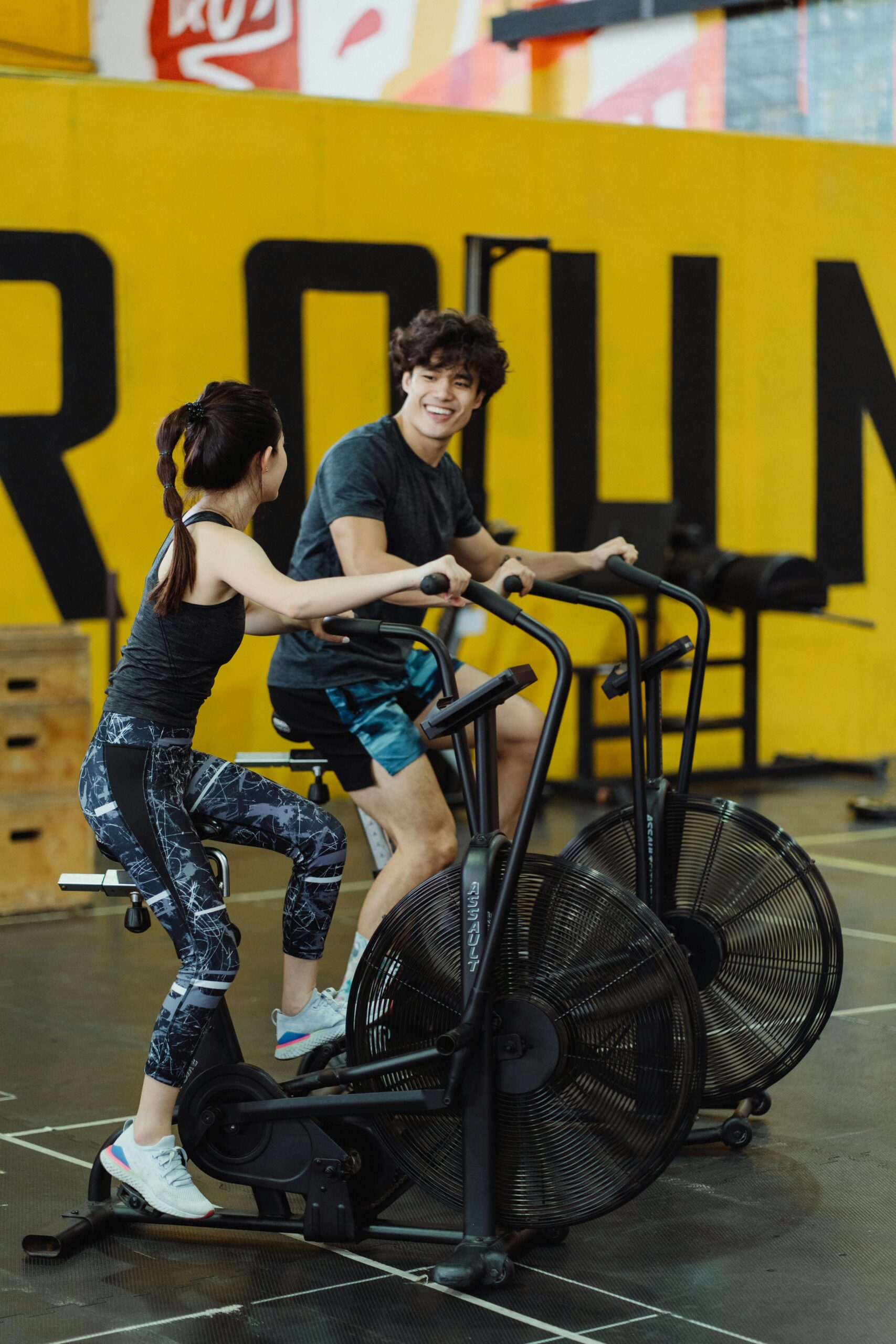 Asian couple on exercise bikes smiling during a gym workout session.