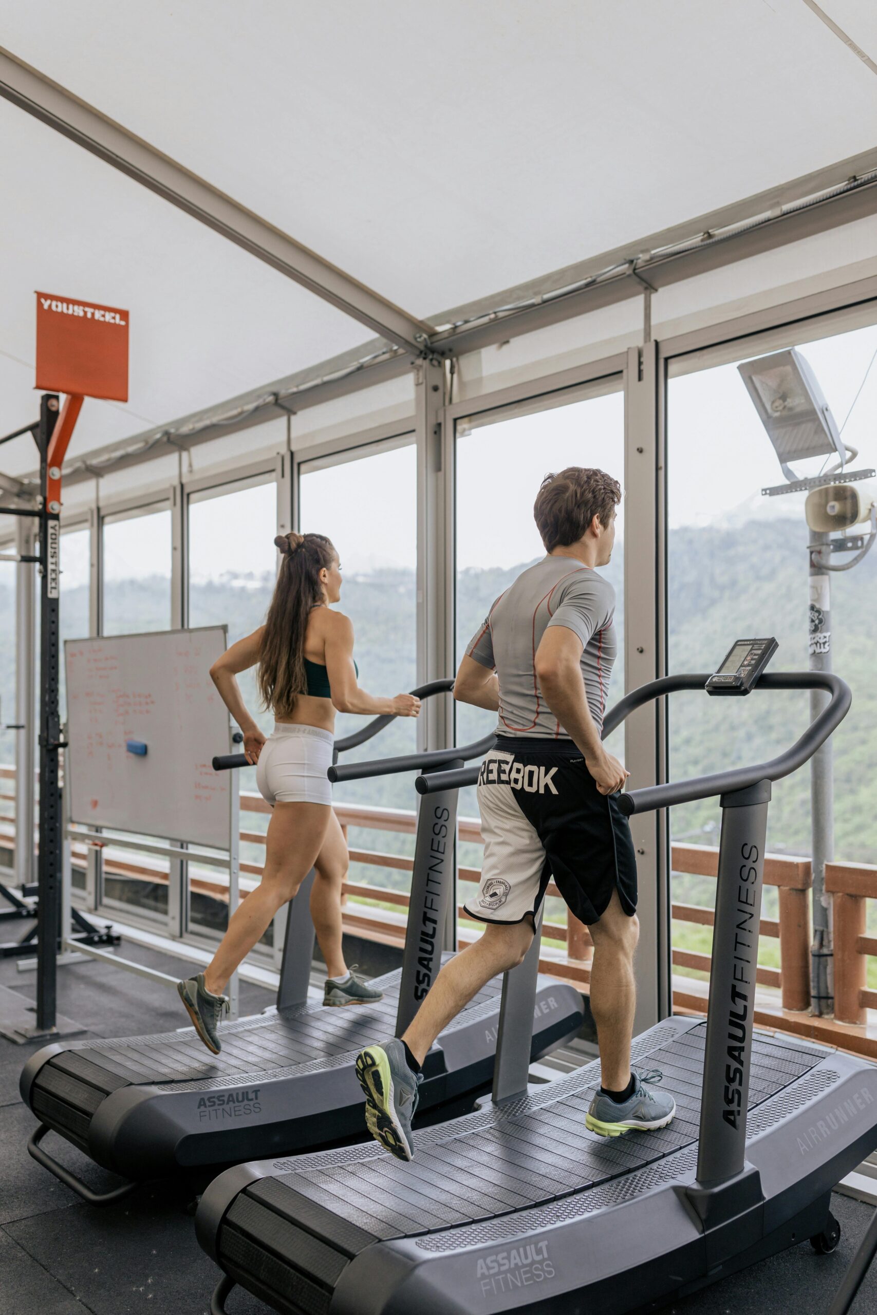 Man and woman running on treadmills in a bright gym with scenic views.