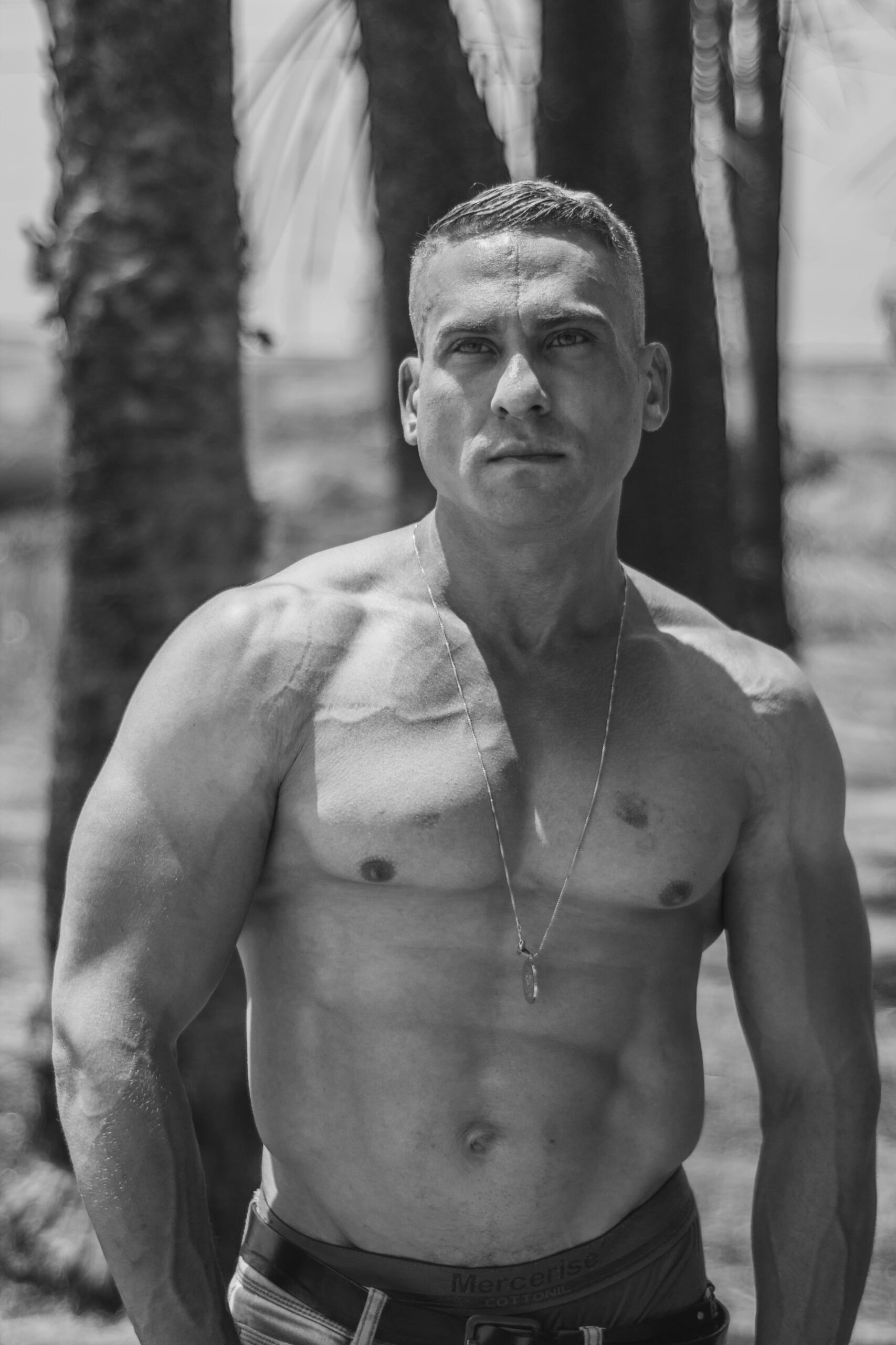 A muscular shirtless man standing outdoors in black and white, showcasing strength.