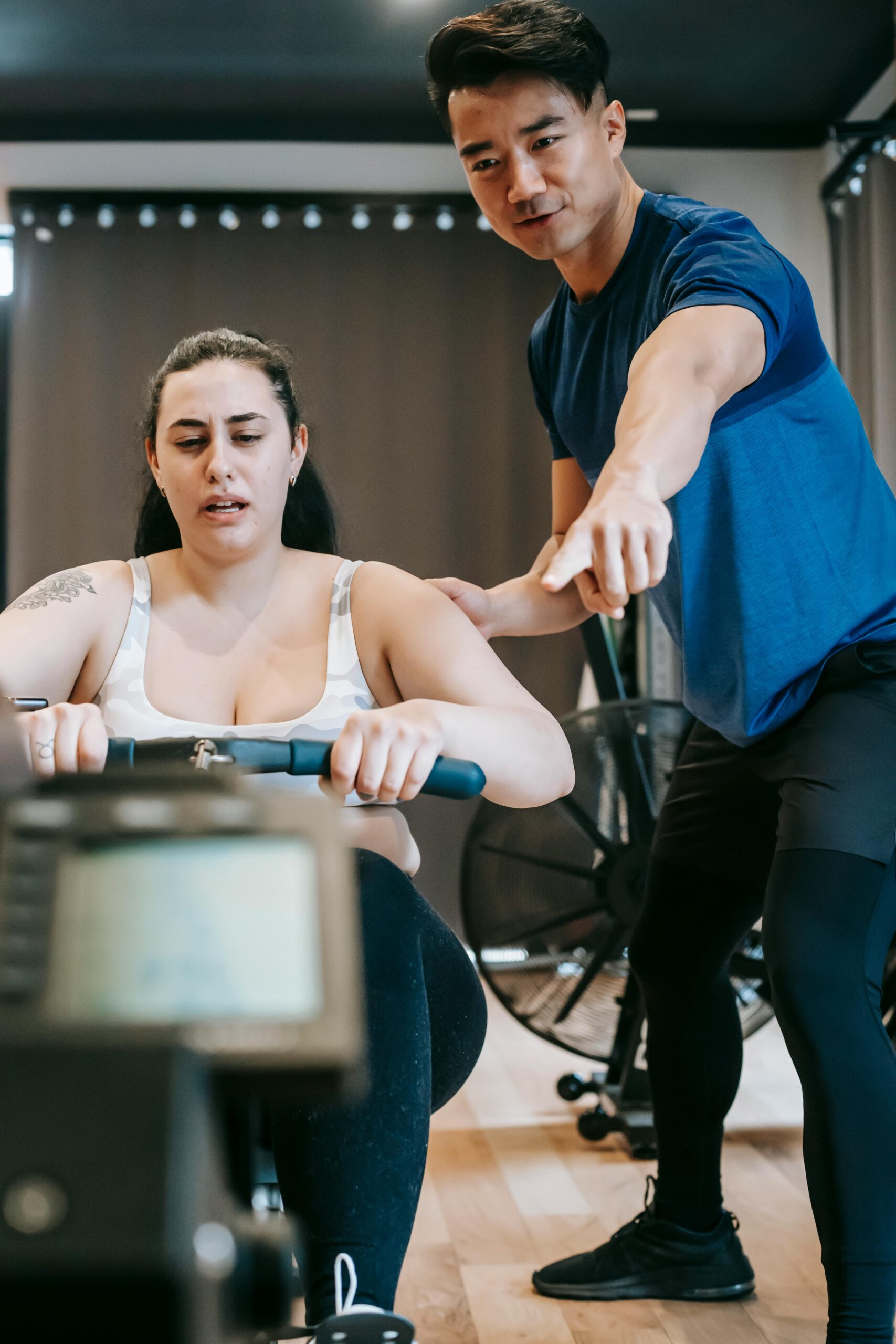 Personal trainer aids woman on rowing machine in gym for effective workout.