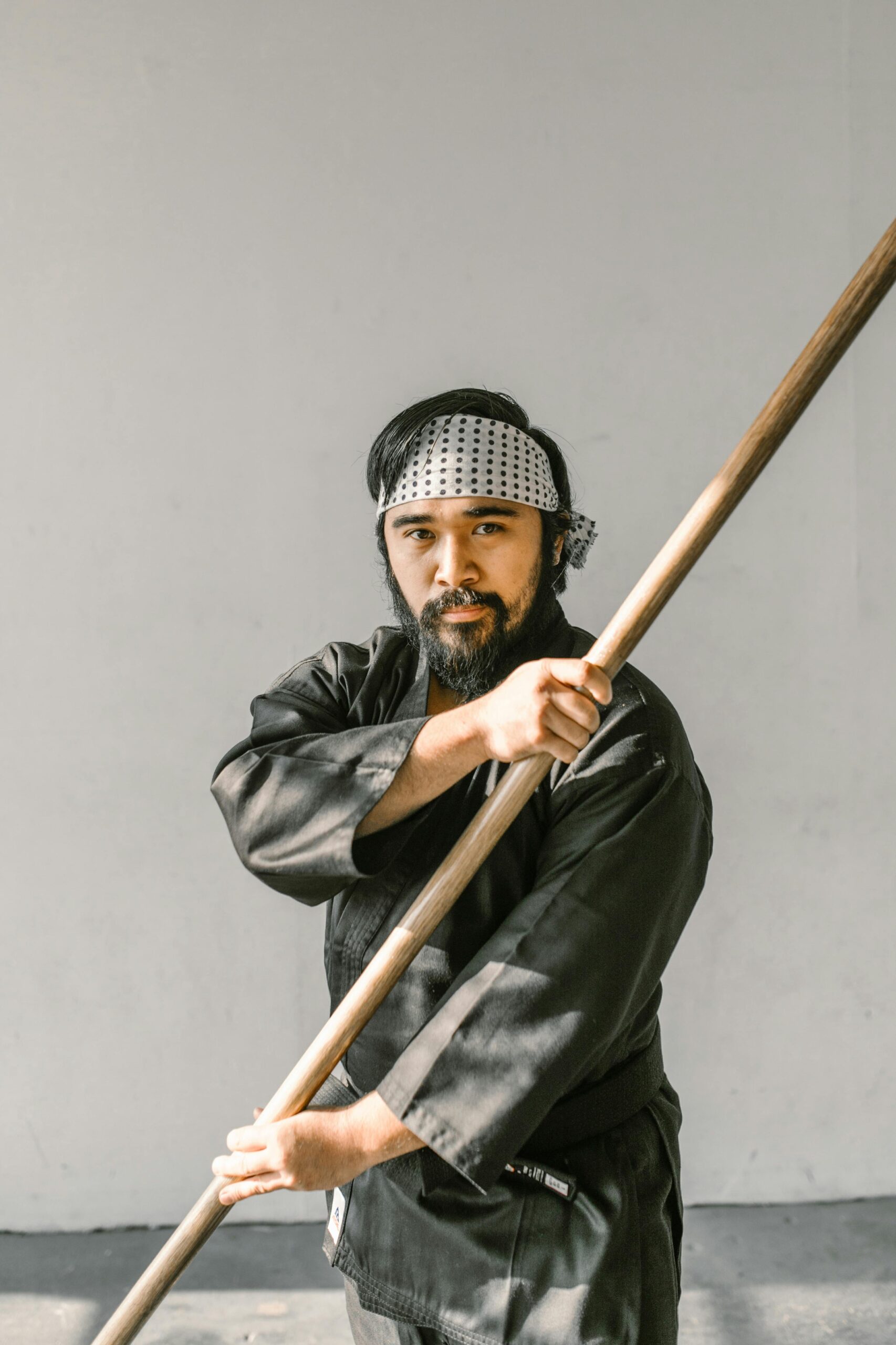 A martial artist demonstrating a pose with a bo staff, indoors against a minimalist background.