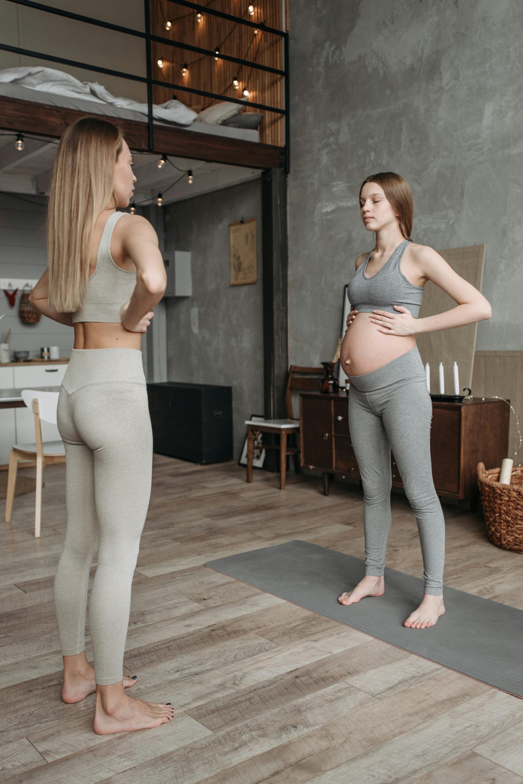 A pregnant woman practices yoga in a cozy indoor setting with a trainer. Promotes wellness and fitness.