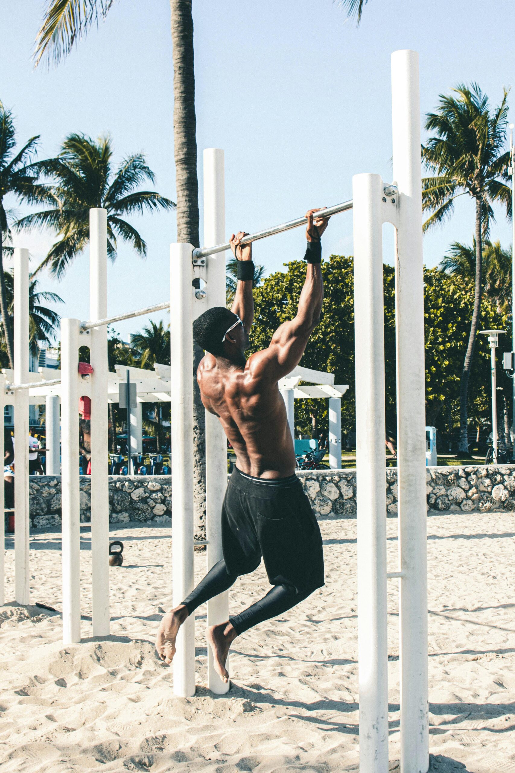 Young man doing pull-ups on a beach gym with palm trees in the background.